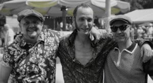 Drie mannen staan buiten met hun armen om elkaar heen en poseren voor de camera. Ze dragen casual shirts met patronen en hoeden. Op de achtergrond zie je een menigte en een groot afdak.