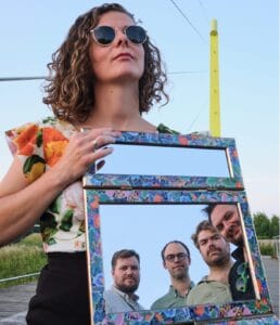 Een vrouw met een zonnebril houdt een versierde spiegel vast waarin de gezichten van vier mannen te zien zijn die buiten onder een heldere hemel staan.