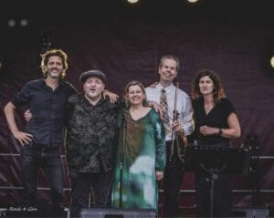 Vijf volwassenen staan dicht bij elkaar op een podium en glimlachen naar de camera. Eén van hen houdt een viool vast en een ander staat naast een muziekstandaard. Achter hen zijn podiumverlichting en een donkere achtergrond te zien.