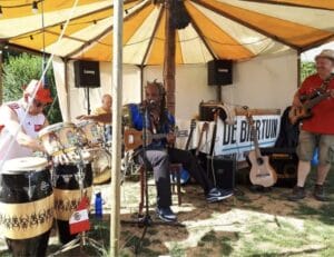 Een vierkoppige band treedt buiten op onder een tent; één van hen speelt gitaar en zingt, de anderen spelen bas, drums en percussie. Rondom hen staan instrumenten en luidsprekers.