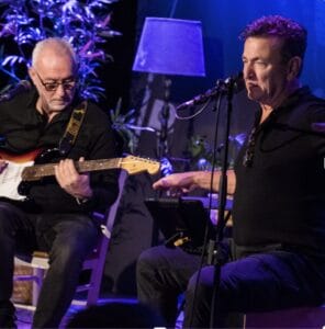Twee mannen treden op het podium op; de ene speelt zittend op een elektrische gitaar, de andere zingt in een microfoon, beiden in het licht van de podiumverlichting, met planten en een lamp op de achtergrond.