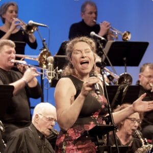 Een vrouw zingt op het podium in een microfoon, omringd door zittende en staande muzikanten die koperblazers bespelen.