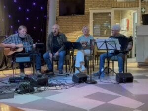 Vier mensen zitten op een podium binnen en bespelen instrumenten – gitaar, microfoon, ukelele en accordeon – met muziekstandaards en geluidsapparatuur.