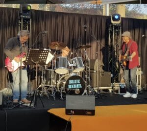 Drie muzikanten van de band Blue Rebel treden op een openluchtpodium op, waar ze elektrische gitaar, drums en bas spelen tegen een achtergrond van zwarte gordijnen en podiumapparatuur.