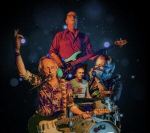 Vier mannelijke muzikanten treden op het podium op met gitaren, bas, drums en mondharmonica, tegen een donkere achtergrond met hier en daar lichtvlekken.