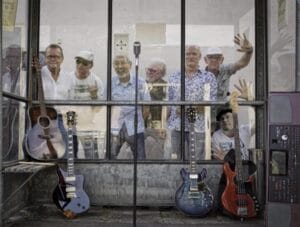 Zeven oudere mannen staan achter een glazen wand, met gitaren, een basgitaar en een keyboard die voor hen buiten zijn neergezet, en een vintage microfoon in het midden.