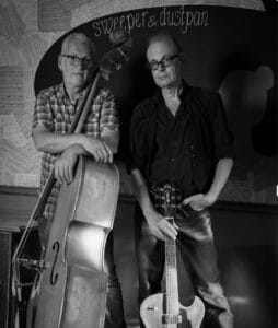Twee mannen staan binnen met muziekinstrumenten; de ene houdt een contrabas vast en de andere heeft zijn hand op een elektrische gitaar gelegd. Aan de muur achter hen hangen bladmuziekpartituren.