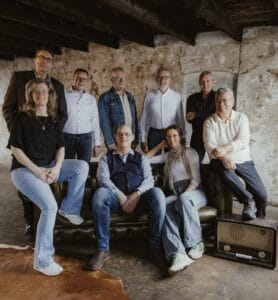 Negen volwassenen poseren voor een groepsfoto in een rustieke kamer met stenen muren; sommigen zitten op een bank, terwijl anderen eromheen staan, naast een oude radio.