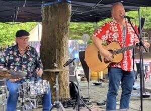 Twee mannen treden buiten op; de ene speelt drums en draagt een zwarte hoed en een bloemenhemd, terwijl de andere zingt en akoestische gitaar speelt, gekleed in een rood bloemenhemd.
