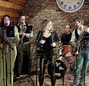 Een vijfkoppige band treedt binnenshuis op; drie leden bespelen instrumenten, terwijl twee anderen zingen. Op de achtergrond zijn een bakstenen muur en een grote klok te zien.