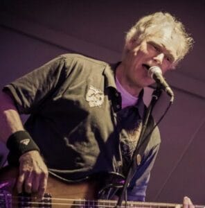 Een man met grijs haar die op het podium gitaar speelt en in een microfoon zingt, gekleed in een donker shirt en met een polsbandje om.