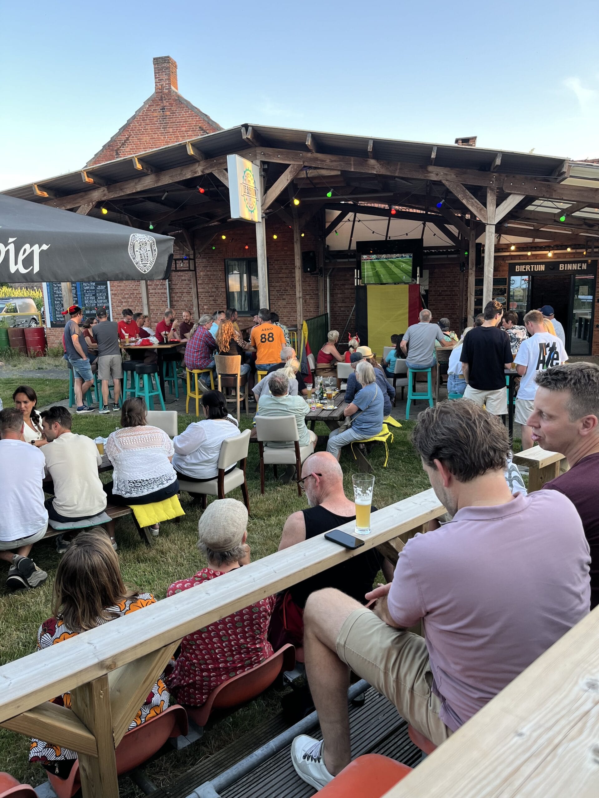 Mensen kijken naar een sportwedstrijd op een buiten-tv in een bar; ze zitten op bankjes en stoelen onder een houten luifel, met drankjes en eten op de tafels.